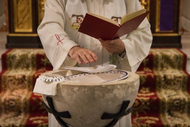 The priest blesses with his hands the water of the baptismal font to proceed to the sacrament of baptism in a catholic church. On the edge of the font the silver shell and the white handkerchief clipart