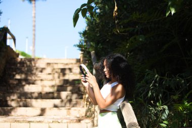 Genç, güzel, Latin, kıvırcık saçlı, spor giysili bir kadın, eğitimden sonra merdivenlerin altındaki ahşap bir çitte dinlenmek için kulaklıklarını çıkarıyor.