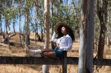 Kadın, genç, güzel, Latin, kıvırcık saçlı, beyaz gömlekli, kot etekli, beyaz çizmeli ve şapkalı, bacakları okaliptüs ormanının çitlerinde uzanmış oturuyor.