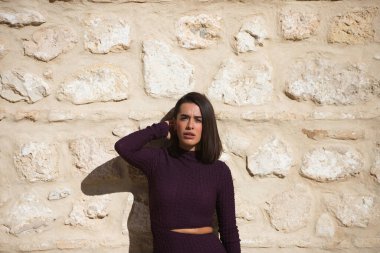 Zarif, menekşe bir elbise giymiş, taş bir duvarda poz veren güzel bir kadın. Kadının durumu ciddi, güneş yüzüne vuruyor, elini başına koyuyor ve önüne bakıyor.