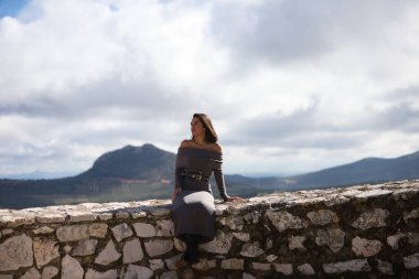 Genç ve güzel bir kadın, esmer, kısa saçlı, gri giysili, siyah çizmeli ve deri kemerli taş duvarda oturuyor, arka planda Hintli yüzüyle dağlarda.