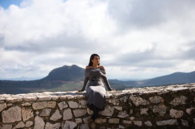 Genç ve güzel bir kadın, esmer, kısa saçlı, gri giysili, siyah çizmeli ve deri kemerli taş duvarda oturuyor, arka planda Hintli yüzüyle dağlarda.