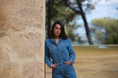 Genç ve güzel Latin kadın, kısa saçlı esmer, kot tulum giymiş Seville, İspanya 'da eski bir manastırın taş duvarına yaslanmış.