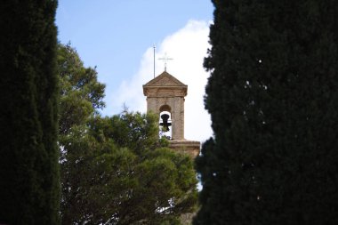 Üzerinde bronz çan olan kilise kulesi, üzerinde dövülmüş demir bir haç var. Arka planda mavi gökyüzü ve beyaz bulutlar