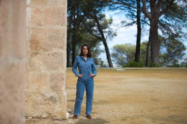 Genç ve güzel Latin kadın, kısa saçlı esmer, kot tulum giymiş Seville, İspanya 'da eski bir manastırın taş duvarına yaslanmış.