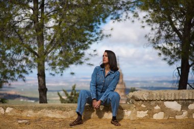 Latin bir kadın, genç ve güzel, taş bir duvarda oturuyor. Arka planda, mavi gökyüzü ve beyaz bulutlardan oluşan arka planda şehir ve kilise kulesini görebilirsiniz.