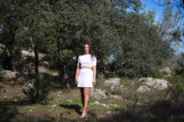 Güzel esmer Latin kadın parkta ağaçların arasında yürürken farklı yüz ve vücut hareketleri yapıyor ve eğleniyor. Güneşli bir sonbahar günü.