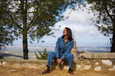 Latin bir kadın, genç ve güzel, taş bir duvarda oturuyor. Arka planda, mavi gökyüzü ve beyaz bulutlardan oluşan arka planda şehir ve kilise kulesini görebilirsiniz.