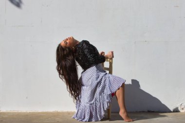 Latin kadın, genç ve güzel, dans eden flamenko, eski tahta bir sandalyede oturuyor ve kafasını geriye atıyor. Kadın, siyah benekli ve siyah ceketli, beyaz, dalgalı bir elbise giyiyor.