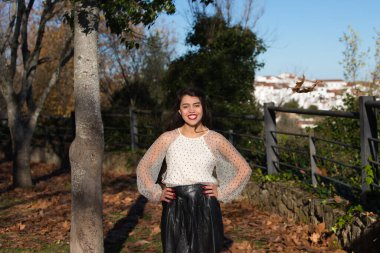 Latin kadın, genç ve esmer makyajlı ve beyaz şeffaf gömlekli siyah benekli ve siyah etekli. Sonbaharda bir parkta, kuru yapraklarla kaplı.