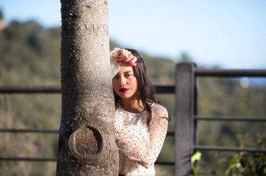 Latin bir kadın, genç ve esmer, makyajlı ve şeffaf beyaz bir gömlek giyiyor siyah benekli ve siyah etekli başını ağaçtaki bir kola yaslıyor. Genç kadın depresyon ve üzüntü içinde.