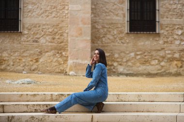 Güzel Latin bir kadın, kısa saçlı esmer, kot tulum giymiş, Seville, İspanya 'da eski bir manastırın yanındaki merdivende oturuyor.