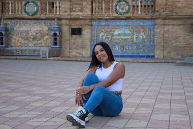 Faslı esmer kadın, genç ve güzel, beyaz bluzlu ve kot pantolonlu, yerde oturuyor ve kollarıyla bacağını tutuyor, kız Seville 'in kare İspanya' sı. Seyahat ve turizm kavramı