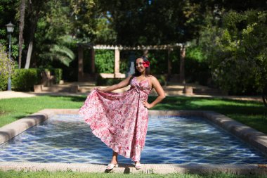 Genç, güzel esmer kadın, uzun çiçekli elbisesi ve saçında çiçek deseni var. Kadın elbisesini eliyle kaldırır ve gülümser. Tatil ve seyahat kavramı