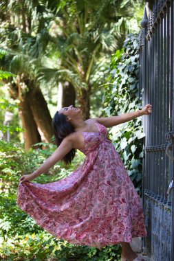 Uzun çiçek desenli elbisesi içindeki genç, güzel esmer kadın elleriyle çiti tutuyor ve vücudunun geriye doğru düşmesine ve elbisesini tutmasına izin veriyor. Kadın mutlu ve eğleniyor.
