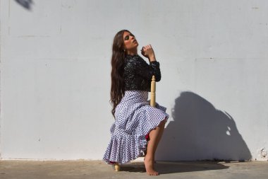 Latin bir kadın, genç ve güzel, dans eden flamenko, eski bir tahta sandalyede oturmuş kameraya bakıyor. Kadın, siyah benekli ve siyah ceketli, beyaz, dalgalı bir elbise giyiyor.