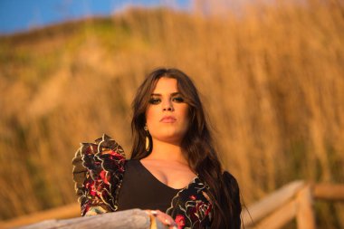 İspanyol kadın, esmer ve güzel, tipik flamenko dans kostümlü dans eden flamenko, tahta korkuluklara yaslanmış ve gün batımında kameraya bakıyor. Kadın üzgün.