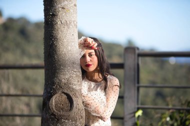 Latin bir kadın, genç ve esmer, makyajlı ve şeffaf beyaz bir gömlek giyiyor siyah benekli ve siyah etekli başını ağaçtaki bir kola yaslıyor. Genç kadın depresyon ve üzüntü içinde.