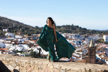 Latin brunette woman, young and beautiful is dressed in green princess dress very elegant on the stone wall of the castle. In the background the village of cortegana in huelva, andalusia, Spain clipart