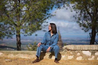 Latin bir kadın, genç ve güzel, taş bir duvarda oturuyor. Arka planda, mavi gökyüzü ve beyaz bulutlardan oluşan arka planda şehir ve kilise kulesini görebilirsiniz.