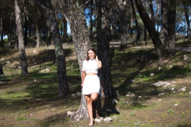 Beyaz elbiseli, güzel esmer kadın. Sırtı parktaki bir ağaca dönük. Kadın mutlu, gülümsüyor ve güneşli sonbahar gününün tadını çıkarıyor. Sarı gün konsepti