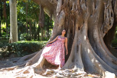 Seville 'deki parktaki dev bir incir ağacının köklerine dayanan çiçek desenli uzun elbiseli güzel esmer kadın. Kadın mutlu, rahat ve sakin.