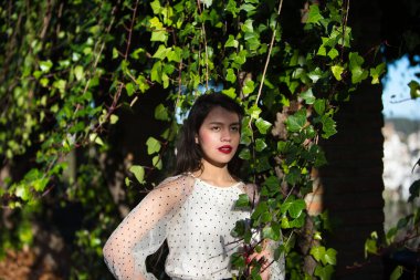 Latin bir kadın, genç ve esmer bir kadın şeffaf beyaz bir gömlek giyip sarmaşıkların arasında siyah benekli bir sarmaşık giyiyor. Kadın mutlu ve eğleniyor.