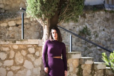 Zarif, menekşe elbiseli genç bir kadın parkın merdivenlerindeki selvi ağacında poz veriyor. Kadın ciddi, yüzünde güneş var ve farklı ifadeler takınıyor.