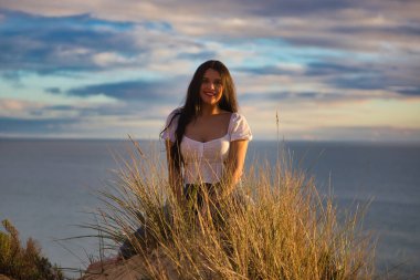 Genç ve güzel Latin kadın, esmer, sazlıkların arasında beyaz bir kum tepesinde oturuyor. Kadın mutlu ve gülümsüyor. Arka planda deniz ve bulutlu mavi gökyüzü.