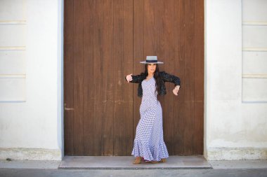 Latin bir kadın, Endülüs 'ün tipik bir sokağındaki ahşap bir kapının önünde dans eden genç ve güzel bir flamenko. Kadın, siyah benekli, şapkalı ve ceketli beyaz bir elbise giyiyor.