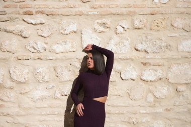 Zarif, menekşe bir elbise giymiş, taş bir duvarda poz veren güzel bir kadın. Kadının yüzünde güneş var ve bir kolunu başının üstüne kaldırıyor ve yana bakıyor.