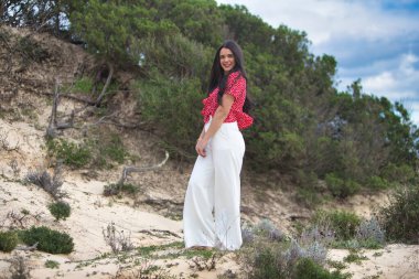 Kumsalın oradaki kumullarda çıplak ayaklı genç, esmer kadın. Kadın gülümsüyor ve mutlu. Kadın beyaz pantolon ve kırmızı gömlek giyiyor.