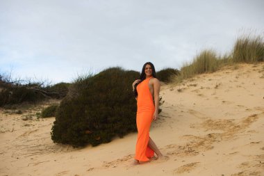 Zarif turuncu elbiseli genç ve güzel esmer kadın İspanya 'nın Endülüs sahilindeki güzel kumulların arasında yürüyor. Kadın farklı duruşlar sergiliyor.