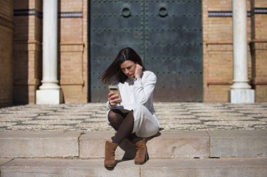 Genç ve güzel Latin esmer kadın merdivenlerde oturmuş cep telefonuna bakıyor. Kadın dikkatle cep telefonu ekranına bakıyor.