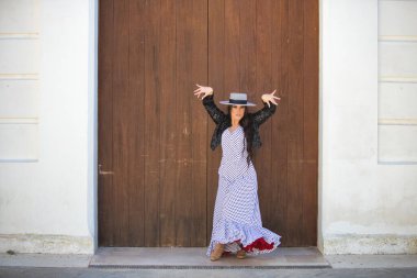 Latin bir kadın, Endülüs 'ün tipik bir sokağındaki ahşap bir kapının önünde dans eden genç ve güzel bir flamenko. Kadın, siyah benekli, şapkalı ve ceketli beyaz bir elbise giyiyor.