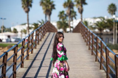 Latin kadın, esmer, dalgalı saçlı, deniz kıyısında tahta bir podyumda flamenko dansı yapan. Kadın çiçek desenli ve fırfırlı beyaz bir elbise giyiyor. Soyut Kültür Mirası