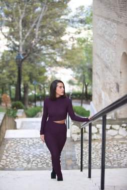 Zarif mor elbiseli güzel bir kadın parkın merdivenlerinden çıkıp eliyle metal korkuluklara tutunuyor. Kadın farklı ifadeler ve jestler yapıyor.