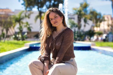 Genç, güzel sarışın kadın parkta dikdörtgen bir fıskiyenin kenarında oturuyor. Kadın mutlu ve gülümsüyor.