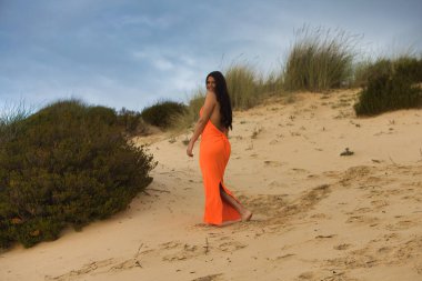 Zarif turuncu elbiseli genç ve güzel esmer kadın İspanya 'nın Endülüs sahilindeki güzel kumulların arasında yürüyor. Kadın farklı duruşlar sergiliyor.