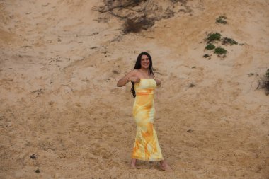 Sarı elbiseli, genç, güzel, yalın ayaklı esmer kadın düşmesin diye elbisesini yukarı çekiyor. Kadın mutlu ve gülümsüyor. Beyaz kumların üzerinde yürürken.