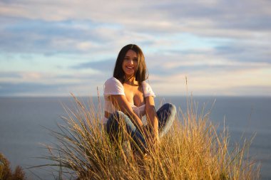 Genç ve güzel Latin kadın, esmer, sazlıkların arasında beyaz bir kum tepesinde oturuyor. Kadın mutlu ve gülümsüyor. Arka planda deniz ve bulutlu mavi gökyüzü.