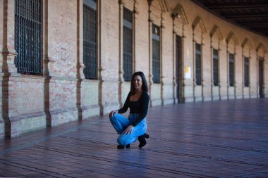 Kot pantolon ve siyah tulum giymiş güzel esmer kadın Seville, Endülüs 'teki İspanya' nın ünlü meydanında çömeliyor. Kadınlar anıtın tadını çıkarıyor.