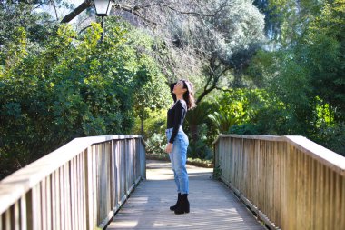 Gündelik giysiler içinde genç, güzel esmer kadın tahta bir köprünün üzerinde yürüyor. Kadın farklı vücut duruşları sergiliyor. Arka planda yeşil bitki örtüsü