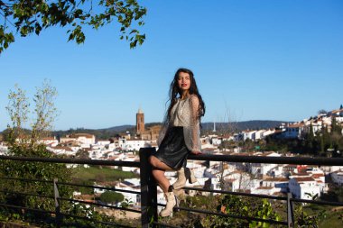 Latin kadın, genç ve esmer Endülüs 'te bir köyde çitin üzerinde oturuyor. Arka planda Beyaz Saray köyü ve kilise. Kadın makyajlı.