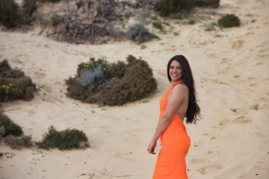 Zarif turuncu elbiseli genç ve güzel esmer kadın İspanya 'nın Endülüs sahilindeki güzel kumulların arasında yürüyor. Kadın farklı duruşlar sergiliyor.