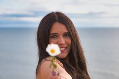 Genç, güzel esmer bir kadının portresi elinde beyaz bir çiçek tutup kameraya gösteriyor. Kız mutlu ve gülümsüyor. Arka planda gökyüzünün denizle buluştuğu ufuk.