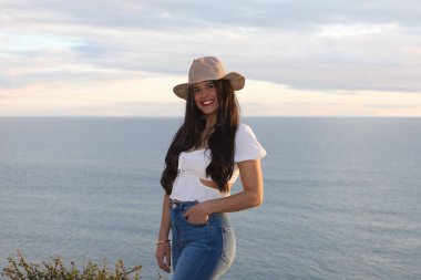 Latin kadın, esmer, genç ve güzel ince beyaz kumdan bir uçurumda. O, kot pantolon ve hasır şapka giydi. Kadın gülümser ve gün batımında altın saatinde kameraya bakar.