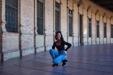 Kot pantolon ve siyah tulum giymiş güzel esmer kadın Seville, Endülüs 'teki İspanya' nın ünlü meydanında çömeliyor. Kadınlar anıtın tadını çıkarıyor.