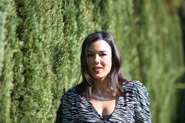 Yolu ayıran yeşil çitlerin önünde duran genç, güzel Latin esmer bir kadının portresi. Kadın mutlu, yolda farklı ifadeler kullanıyor ve eğleniyor.