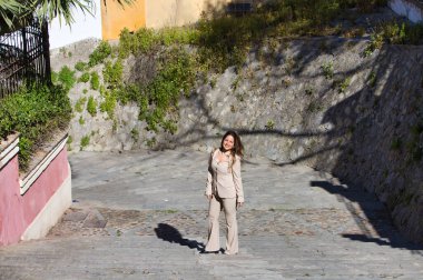 Genç ve güzel sarışın kadın bir caddenin merdivenlerinden yukarı çıkıyor. Kadın bej bir ceket giyiyor. Kadın mutlu ve farklı vücut duruşları sergiliyor. Fotoğraf yukarıdan çekildi